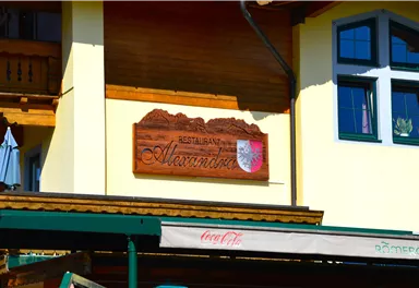 Ein Restaurant mit dem Namen "Alexandra" wird durch ein Holzschild angezeigt. Im Hintergrund sind Teile eines gelben Gebäudes und Fenster zu sehen.