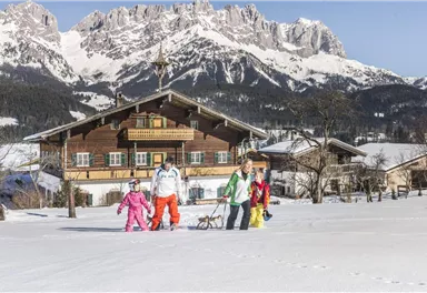 Eine verschneite Landschaft mit einem traditionellen Holzhaus und majestätischen Bergen im Hintergrund. Eine Familie in bunten Skianzügen genießt die winterliche Umgebung.