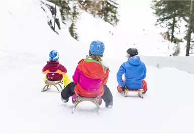 Drei Kinder sitzen auf Schlitten und schauen den Schneeberg hinunter. Sie tragen bunte Winterkleidung und Helme.