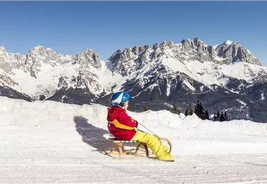 Ein Kind sitzt auf einem Schlitten im Schnee. Im Hintergrund sind beeindruckende Berge zu sehen.
