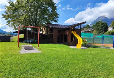 A playground with a slide and swings in the middle of a green meadow. In the background, trees and mountains are visible.