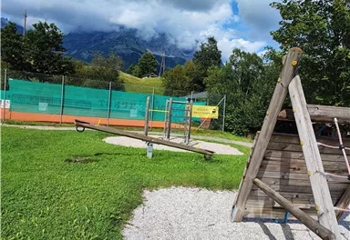 A playground with a seesaw and a wooden frame. In the background, tennis courts and mountains can be seen.