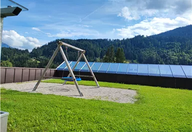 A swing on a sand area in a green garden. In the background, there are hills and trees visible.