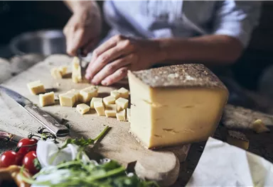 A block of cheese is cut into small pieces on a wooden board. Fresh ingredients like tomatoes and lettuce are in the background.