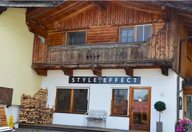 A traditional wooden house with a modern sign "STYLE EFFECT" above the front door. In the foreground, there are wood piles and a small pot with a plant.