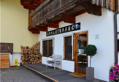 A stylish entrance with the sign "STYLE EFFECT". Next to the door, there is a small table and a plant, while piles of wood can be seen in the background.