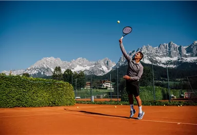 Ein Tennisspieler serviert auf einem Sandplatz. Im Hintergrund sind majestätische Berge zu sehen.