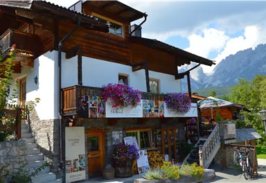 A charming wooden house with a balcony full of flowers. In the background, there are mountains and a clear sky.