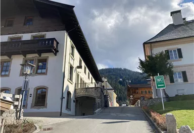 A charming street in the mountains with traditional buildings. In the background, the green hills are visible along with a sign for a restroom.