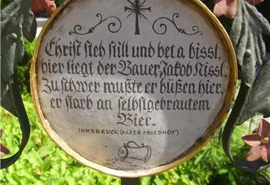 Eine metallene Gedenktafel mit einer Inschrift in alter deutscher Schrift. Die Tafel erinnert an einen verstorbenen Bauer und befindet sich in einem Friedhof.