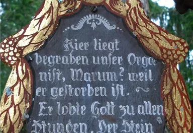 Ein verzierter Grabstein mit einer poetischen Inschrift. Die Umgebung besteht aus Bäumen und einer ruhigen Naturkulisse.