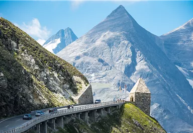 A winding road leads through an impressive mountain landscape. Majestic peaks rise into the clear sky in the background.