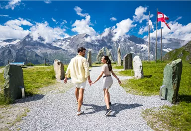 A couple is holding hands and walking on a gravel path. In the background, impressive mountains and clouds are visible.