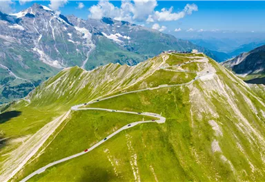 A breathtaking mountain landscape with winding roads and green meadows. In the background, majestic peaks and a clear sky can be seen.