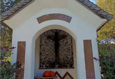 Eine kleine Kapelle mit einem kreuzförmigen Dach und einer schmiedeeisernen Gittertür. Auf einem Tisch vor der Kapelle stehen ein Kürbis und religiöse Symbole.