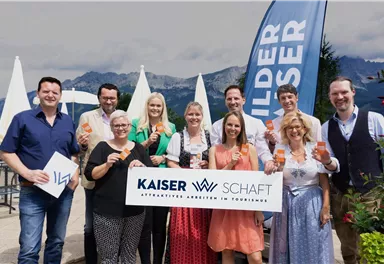 A group of people is standing together, holding drinks in their hands. In the background, mountains and an advertisement banner are visible.