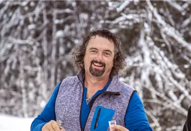 Ein lächelnder Mann im Schnee trägt eine bunte Jacke. Hintergrund ist mit verschneiten Bäumen bedeckt.