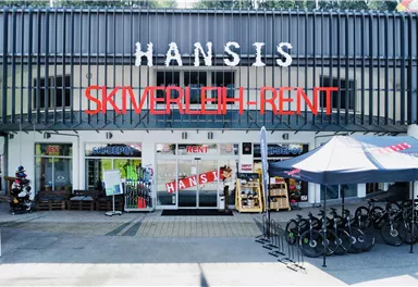 Ein Geschäft mit der Aufschrift „Hansis Skiverleih-Rent“ und einer großen Auswahl an Sportartikeln. Vor dem Geschäft stehen mehrere Fahrräder zur Miete.