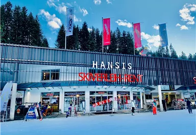 Ein modernes Skigeschäft mit der Aufschrift "HANSIS SKIVERLEIH" inmitten einer verschneiten Umgebung. Im Hintergrund sind Bäume und ein blauer Himmel zu sehen.