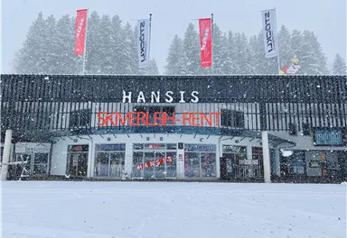 Ein Skiverleih mit dem Namen "Hansis" im Schnee. Die Fassade ist modern, umgeben von schneebedeckten Bäumen.