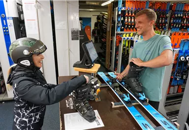 Eine Skifahrerin erhält ihre Ausrüstung in einem Sportgeschäft. Der Mitarbeiter hilft ihr freundlich beim Anlegen der Skischuhe.