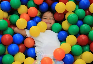 Ein lächelndes Kind liegt in einem bunten Ballbecken. Es ist umgeben von roten, gelben, blauen und grünen Bällen.