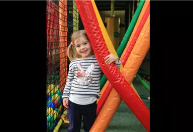 Ein fröhliches Mädchen steht zwischen bunten Spielgeräten in einem Indoor-Spielplatz. Sie lächelt und trägt ein gestreiftes Shirt mit einem Tiermotiv.