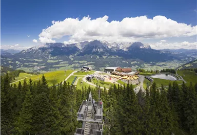 Eine beeindruckende Berglandschaft mit viel Grün und hohen Gipfeln. Im Vordergrund ist eine Bergstation mit gemütlichen Sitzplätzen und Wegen zu sehen.