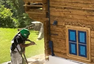 Ein Arbeiter reinigt die Außenwand eines Holzhauses mit einem Hochdruckreiniger. Im Hintergrund sind grüne Wiesen und ein klarer Himmel zu sehen.