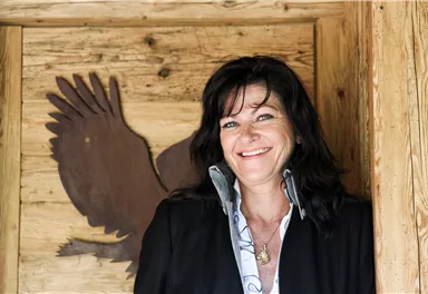 Eine Frau lächelt vor einer hölzernen Wand mit einem Adleremblem. Sie trägt eine elegante Jacke und wirkt freundlich und einladend.