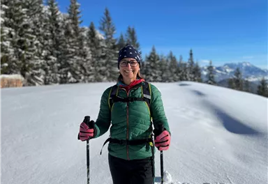 A woman stands joyfully in the snow, surrounded by snow-covered trees. The sky is clear and blue, and she is holding ski poles in her hands.