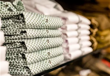 Stacked shirts, some with a green pattern, are stored on a shelf. In the background, more folded shirts are visible.