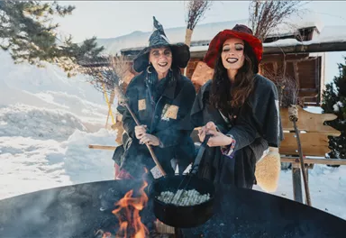 Zwei Frauen in kreativen Hexenkostümen stehen am Feuer und kochen in einem großen Topf. Im Hintergrund ist Schnee zu sehen, was eine winterliche Atmosphäre schafft.