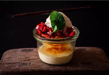 A delicious dessert in a glass, consisting of a cream with a caramelized surface, fresh fruits, and whipped cream. A mint leaf decorates the dish.
