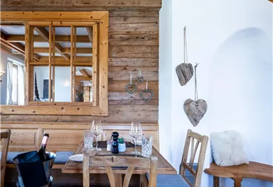 A cozy wooden room with a wooden table and chairs. Wooden decorations hang on the wall, and there is a large window reflection.