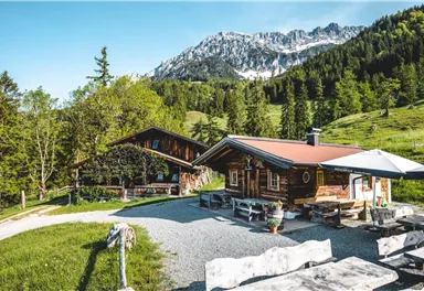 Eine malerische Almhütte umgeben von grünen Wiesen und Bergen. Die Sonne scheint und der Himmel ist klar.