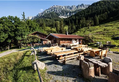 Eine gemütliche Almhütte in einer bergigen Landschaft. Tische und Bänke aus Holz laden zum Verweilen ein.