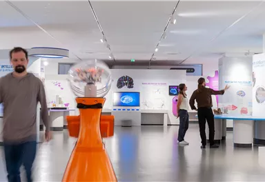 Ein modernes Museum mit interaktiven Ausstellungen. Menschen erkunden verschiedene Stationen in einem hellen und offenen Raum.
