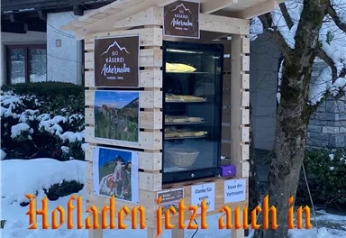 A small outdoor sales booth for local products stands in a snowy environment. The booth displays signs with the name "Käserei Ackerhube" and information about the offerings.