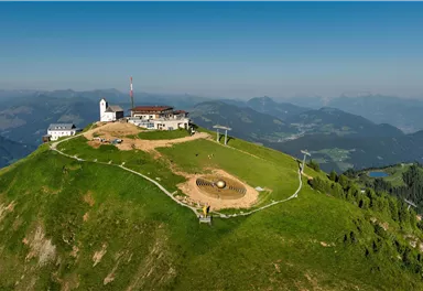 A picturesque mountain landscape with a viewpoint and several buildings. In the background, gentle hills and a clear blue sky can be seen.