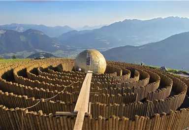 An impressive labyrinth made of wood is situated in a mountainous landscape. In the background, the majestic mountains and a clear sky can be seen.