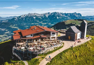 Ein schönes Bergrestaurant auf einem Hügel mit Terrassen und zahlreichen Tischen. Im Hintergrund erheben sich majestätische Berge unter einem klaren blauen Himmel.
