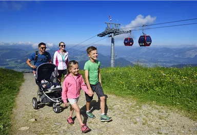 A family enjoys a sunny day in the mountains. In the background, there is a cable car and a blooming landscape.