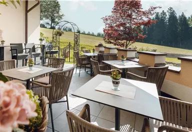 A cozy terrace with tables and chairs, surrounded by green meadows and trees. The fresh air and the flowers create an inviting atmosphere.