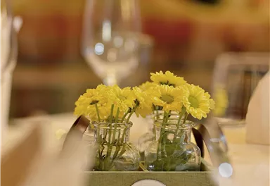 Ein Tischgedeck mit einer kleinen Blumenvase und gelben Blumen. Im Hintergrund sind Gläser und eine dezente Atmosphäre zu sehen.