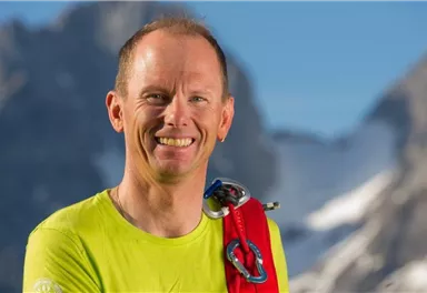 Ein lächelnder Mann in einem leuchtend gelben Shirt steht vor einer bergigen Landschaft. Er trägt Kletterausrüstung und strahlt Abenteuerlust aus.