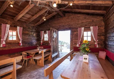 Eine gemütliche Hütte mit Holzwänden und großen Fenstern. Innen sind Holztische und Bänke, sowie dekorative Vorhänge und ein Blumenstrauß.