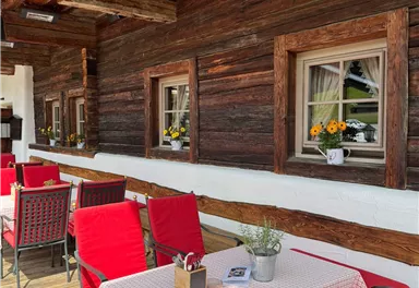 An inviting terrace with red chairs and a table covered with a checkered tablecloth. In the background, the windows of a wooden house can be seen, adorned with flowers.