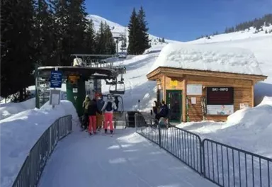 Eine Skipiste mit einer Bergbahn und einem kleinen Informationsgebäude. Rundherum liegt viel Schnee und es ist sonnig.