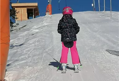 Ein Kind in pinken Skihosen steht auf einer schneebedeckten Piste. Im Hintergrund sind ein Skilift und ein Holzgebäude zu sehen.